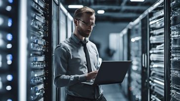 Un homme debout dans une salle de serveurs avec un ordinateur portable.