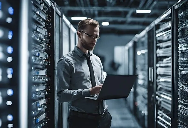 Un homme debout dans une salle de serveurs avec un ordinateur portable.