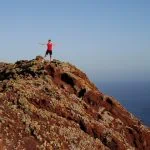 Femme aux bras tendus debout sur une colline rocheuse surplombant l'océan sous un ciel bleu clair, symbolisant la façon dont elle atteint ses objectifs.