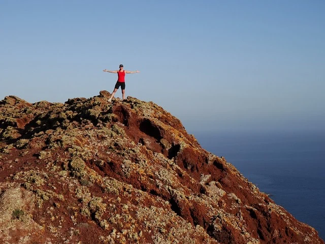 Femme aux bras tendus debout sur une colline rocheuse surplombant l'océan sous un ciel bleu clair, symbolisant la façon dont elle atteint ses objectifs.