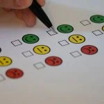 Main tenant un stylo pour cocher une case sur un papier d'enquête avec des icônes colorées de smiley et de visage triste indiquant différents niveaux de satisfaction au travail.