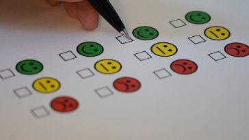 Main tenant un stylo pour cocher une case sur un papier d'enquête avec des icônes colorées de smiley et de visage triste indiquant différents niveaux de satisfaction au travail.