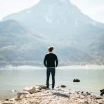 Un homme debout sur une côte rocheuse face à une montagne au-dessus d’un lac tranquille, vivant un moment d’accomplissement personnel.
