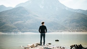 Un homme debout sur une côte rocheuse face à une montagne au-dessus d’un lac tranquille, vivant un moment d’accomplissement personnel.