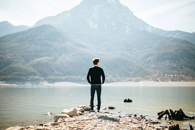 Un homme debout sur une côte rocheuse face à une montagne au-dessus d’un lac tranquille, vivant un moment d’accomplissement personnel.