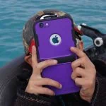 Personne tenant un iPhone violet avec un étui robuste, prenant une photo près de l’eau.