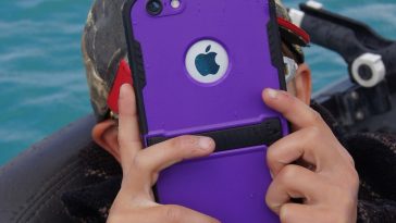 Personne tenant un iPhone violet avec un étui robuste, prenant une photo près de l’eau.