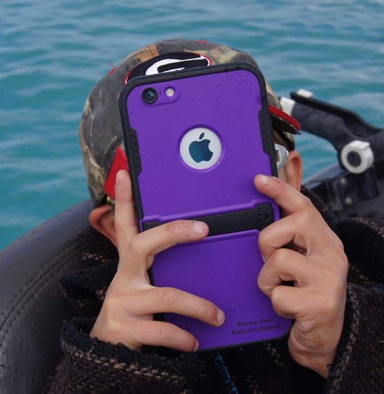 Personne tenant un iPhone violet avec un étui robuste, prenant une photo près de l’eau.