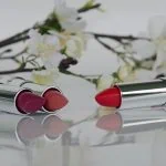 Deux rouges à lèvres, un rose et un rouge, posés horizontalement, capuchons retirés, sur une surface réfléchissante, entourés de fleurs blanches sur fond blanc qui complètent une « routine beauté » quotidienne.