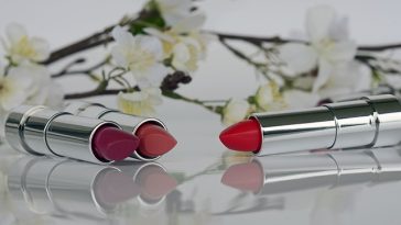 Deux rouges à lèvres, un rose et un rouge, posés horizontalement, capuchons retirés, sur une surface réfléchissante, entourés de fleurs blanches sur fond blanc qui complètent une « routine beauté » quotidienne.