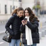 Deux jeunes femmes en vestes d'hiver regardant ensemble un iPhone haut de gamme dans une rue de la ville.