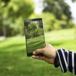Une main tenant un smartphone affichant une photo d'un parc verdoyant, en alignant la photo avec la scène du parc en arrière-plan pour contribuer à la sensibilisation au développement durable.