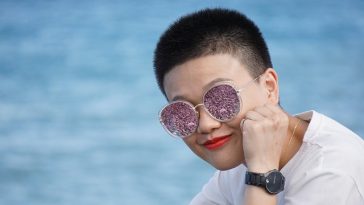 Une personne aux cheveux courts et aux lunettes de soleil violettes scintillantes pose son menton sur sa main, maquillée impeccablement, avec un fond d'eau bleu flou.