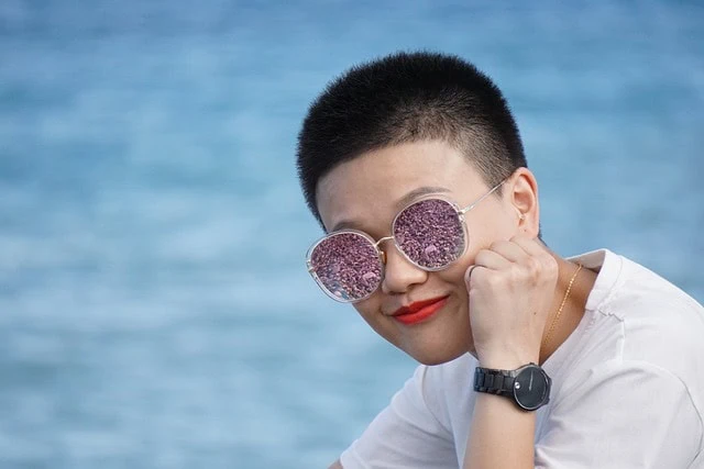 Une personne aux cheveux courts et aux lunettes de soleil violettes scintillantes pose son menton sur sa main, maquillée impeccablement, avec un fond d'eau bleu flou.