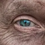 Gros plan sur l'œil d'une personne âgée à l'iris bleu et à la peau ridée, mettant en avant des produits adaptés au vieillissement cutané.