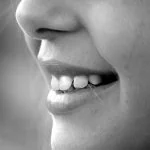 Gros plan de la bouche d'une personne souriante montrant des dents blanches, en noir et blanc, avec un maquillage subtil du nez.
