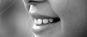 Gros plan de la bouche d'une personne souriante montrant des dents blanches, en noir et blanc, avec un maquillage subtil du nez.