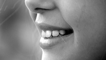 Gros plan de la bouche d'une personne souriante montrant des dents blanches, en noir et blanc, avec un maquillage subtil du nez.