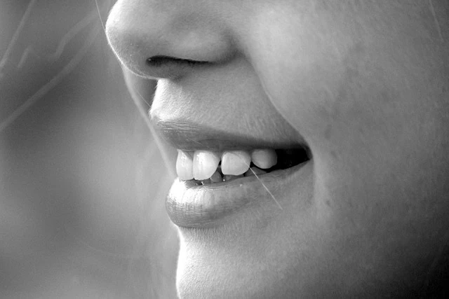 Gros plan de la bouche d'une personne souriante montrant des dents blanches, en noir et blanc, avec un maquillage subtil du nez.