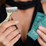 Une personne tenant un rasoir en métal dans une main et trois paquets verts dans l'autre, vêtue d'un haut sans manches noir, prête à choisir la meilleure méthode d'épilation ou à opter pour le rasage maillot.