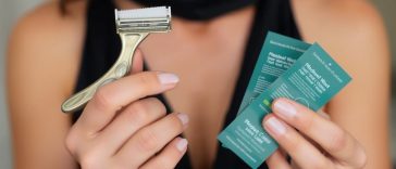 Une personne tenant un rasoir en métal dans une main et trois paquets verts dans l'autre, vêtue d'un haut sans manches noir, prête à choisir la meilleure méthode d'épilation ou à opter pour le rasage maillot.