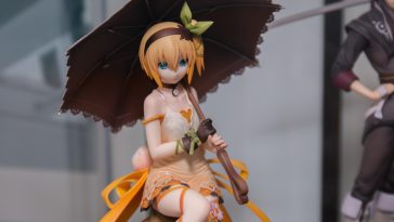 Une figurine geek chic d'une fille de style anime aux cheveux blonds courts, portant une robe crème et tenant un parasol noir, est assise sur le présentoir - un parfait objet de collection pour tout fan.