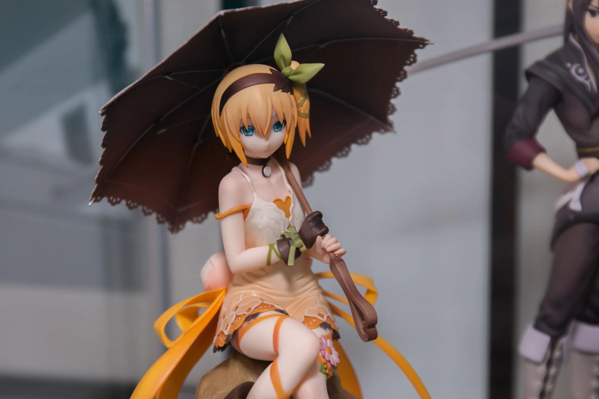 Une figurine geek chic d'une fille de style anime aux cheveux blonds courts, portant une robe crème et tenant un parasol noir, est assise sur le présentoir - un parfait objet de collection pour tout fan.